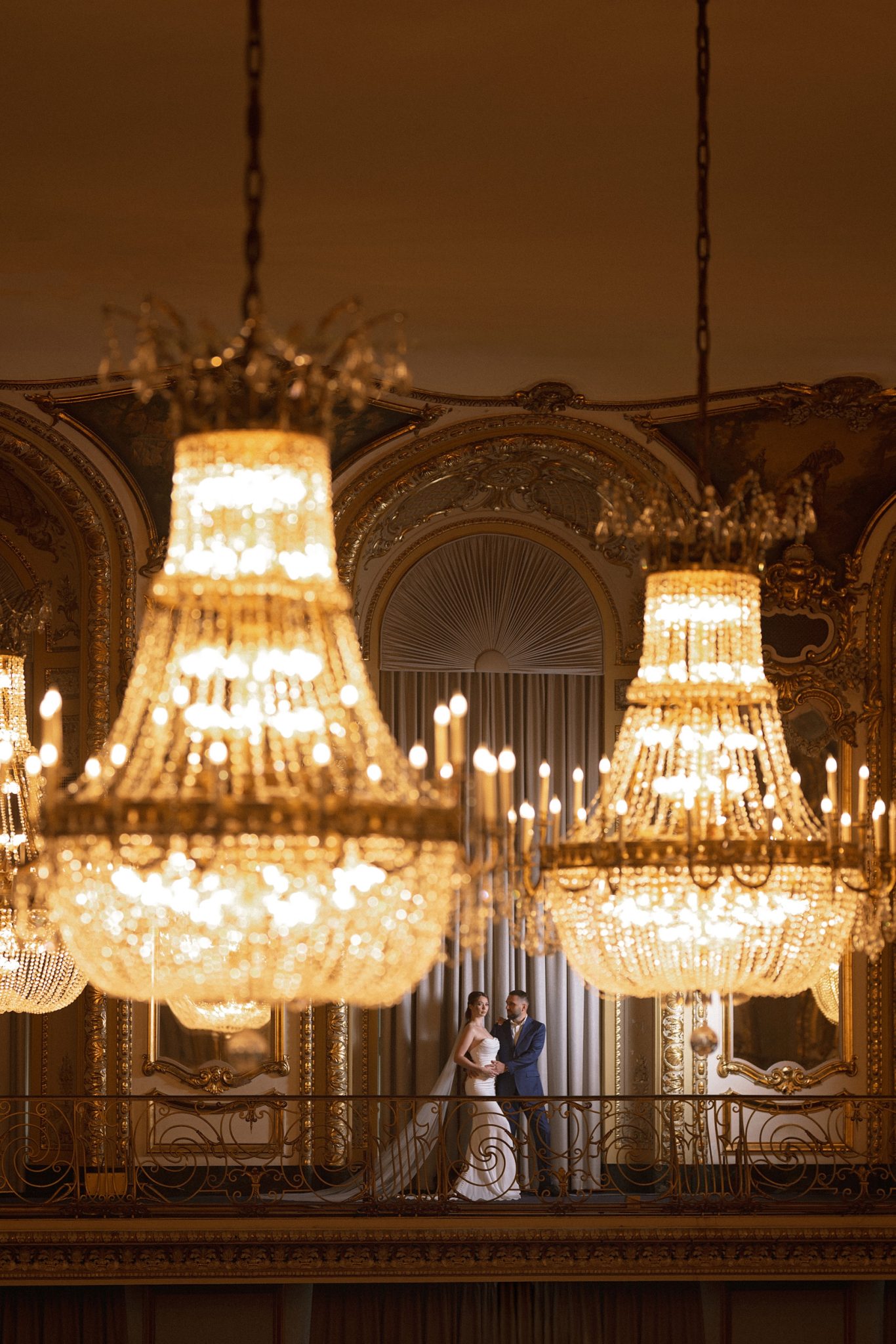 Wide balcony portrait balances the couple with the grand architecture and ambient room light.